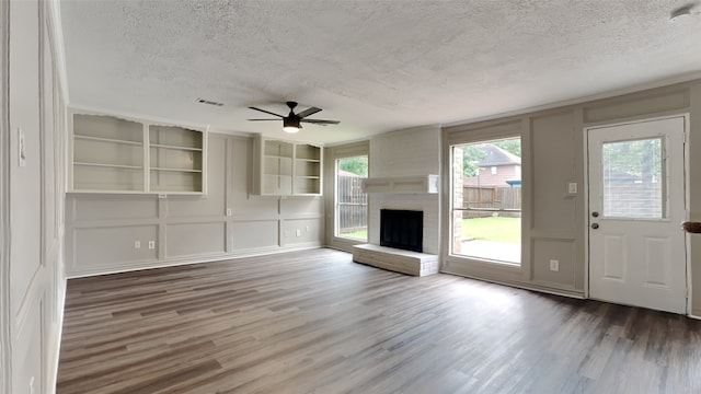 unfurnished living room with wood-type flooring, a brick fireplace, a textured ceiling, built in shelves, and ceiling fan