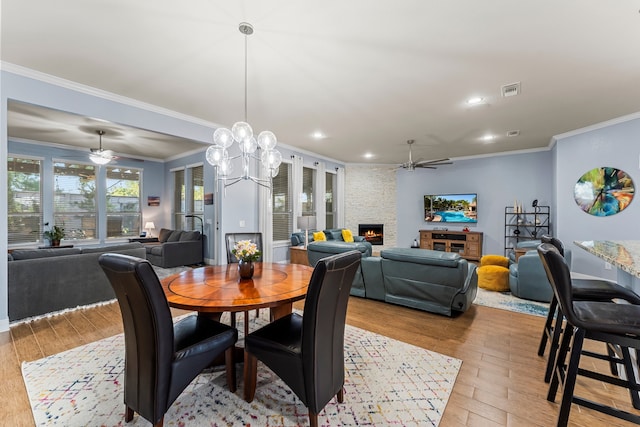 dining room with ceiling fan with notable chandelier, light hardwood / wood-style floors, a stone fireplace, and ornamental molding