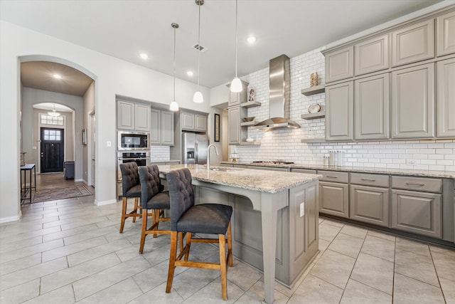 kitchen with light stone countertops, wall chimney range hood, a kitchen bar, stainless steel appliances, and a kitchen island with sink