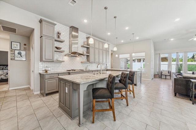 kitchen with decorative backsplash, wall chimney exhaust hood, a breakfast bar area, a center island with sink, and light stone counters