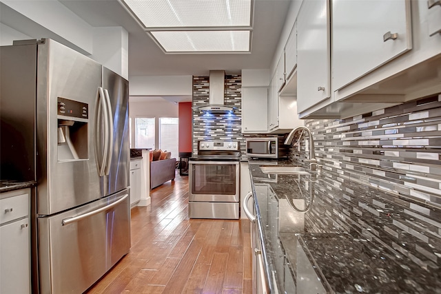 kitchen featuring tasteful backsplash, appliances with stainless steel finishes, wall chimney exhaust hood, white cabinets, and light hardwood / wood-style flooring