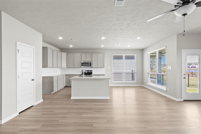 kitchen with light stone countertops, a textured ceiling, gray cabinets, a center island with sink, and light wood-type flooring