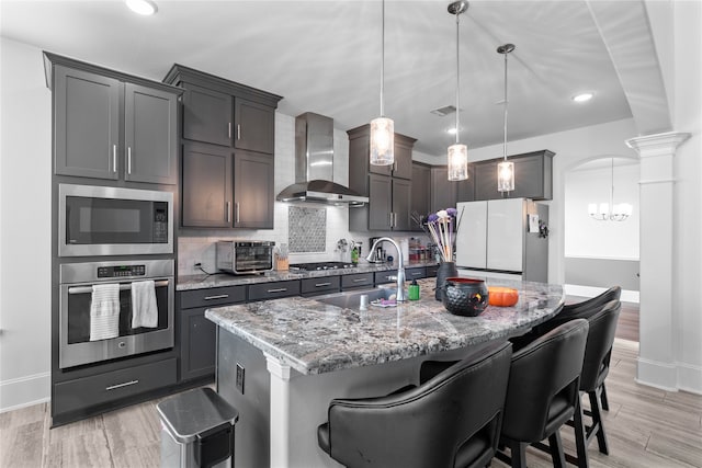 kitchen featuring wall chimney range hood, hanging light fixtures, a center island with sink, a kitchen bar, and appliances with stainless steel finishes