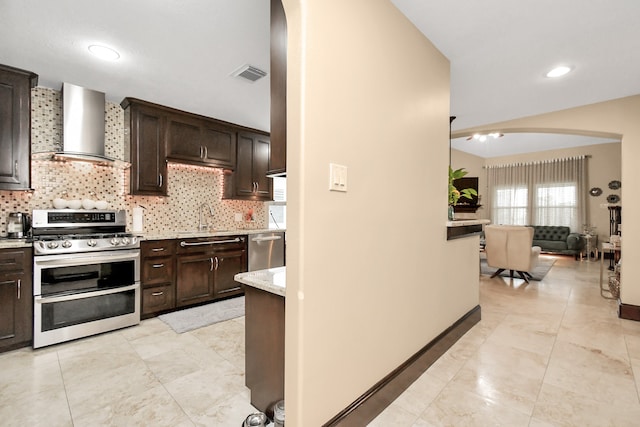 kitchen featuring wall chimney range hood, dark brown cabinets, light stone counters, backsplash, and stainless steel appliances