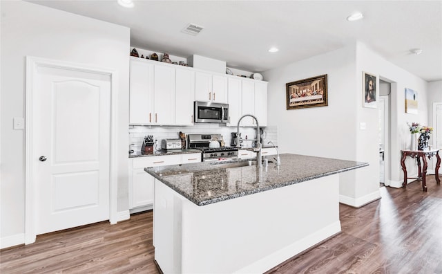 kitchen featuring wood-type flooring, white cabinetry, stainless steel appliances, dark stone counters, and an island with sink