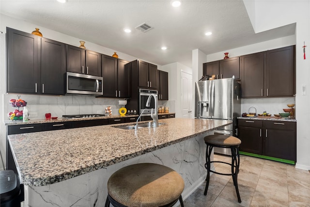 kitchen featuring dark brown cabinets, a center island with sink, a breakfast bar area, appliances with stainless steel finishes, and a textured ceiling