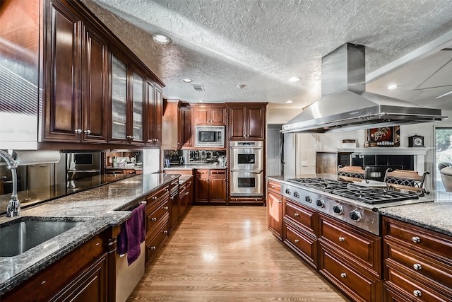 kitchen with island exhaust hood, light hardwood / wood-style flooring, dark stone countertops, sink, and stainless steel appliances