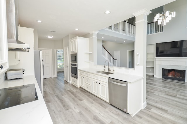 kitchen with sink, appliances with stainless steel finishes, a high end fireplace, white cabinets, and wall chimney range hood