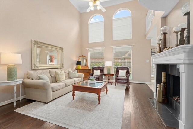 living room with dark wood-type flooring, a towering ceiling, a tiled fireplace, and ceiling fan