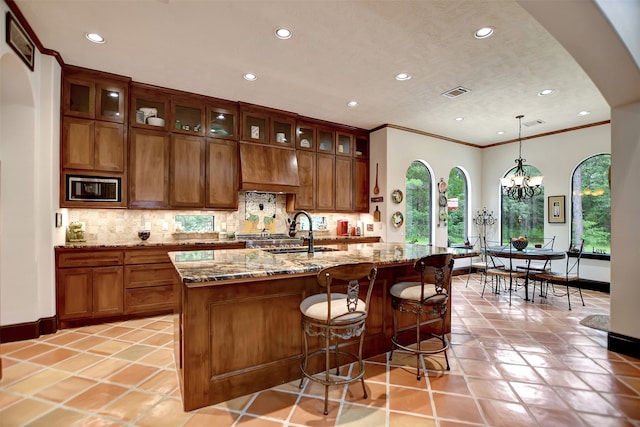 kitchen with a kitchen island with sink, stainless steel microwave, hanging light fixtures, a notable chandelier, and light stone counters