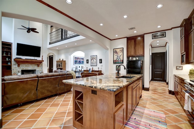 kitchen featuring a stone fireplace, a center island with sink, sink, crown molding, and ceiling fan