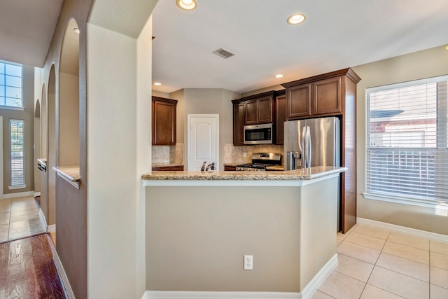 kitchen featuring kitchen peninsula, tasteful backsplash, appliances with stainless steel finishes, light tile patterned flooring, and light stone counters