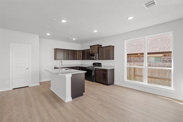 kitchen with sink, light stone countertops, light wood-type flooring, an island with sink, and black range with gas cooktop