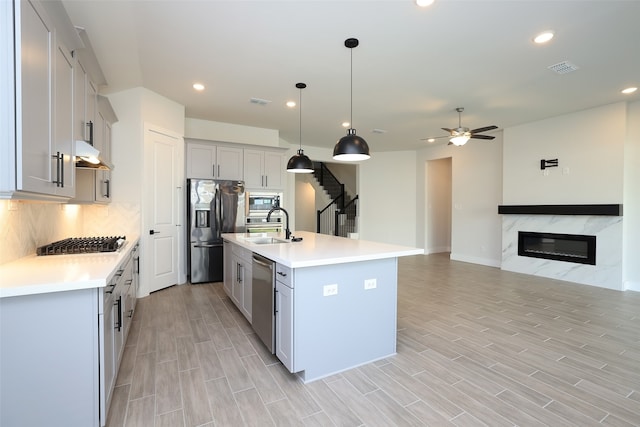 kitchen with exhaust hood, hanging light fixtures, a center island with sink, light wood-type flooring, and stainless steel appliances