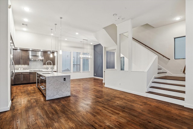 kitchen featuring a kitchen island with sink, hanging light fixtures, dark wood-type flooring, and wall chimney range hood