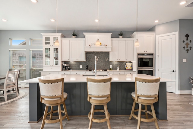 kitchen featuring white cabinets, a center island with sink, hanging light fixtures, light wood-type flooring, and double oven