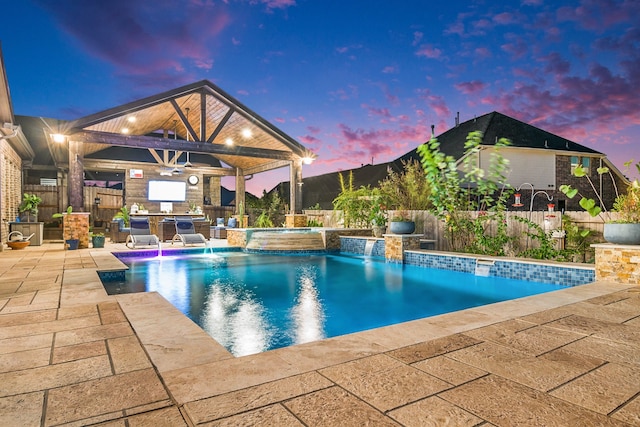 pool at dusk featuring a gazebo, ceiling fan, pool water feature, a patio area, and an in ground hot tub