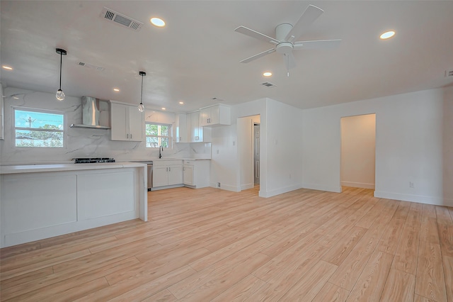 kitchen featuring wall chimney range hood, light hardwood / wood-style flooring, pendant lighting, white cabinetry, and ceiling fan