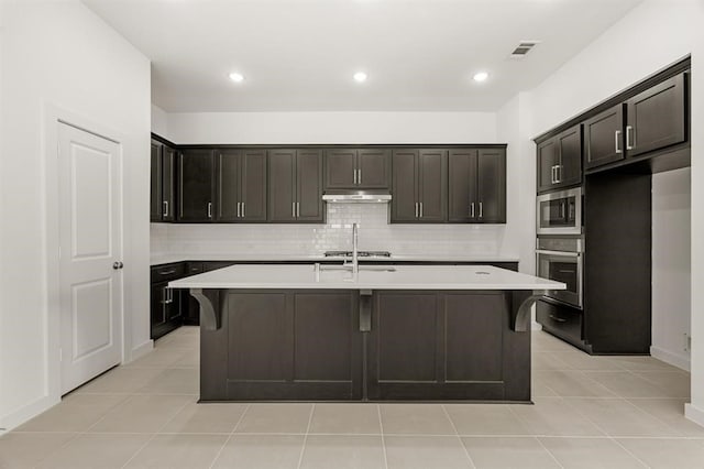 kitchen with light tile patterned flooring, appliances with stainless steel finishes, dark brown cabinetry, and an island with sink