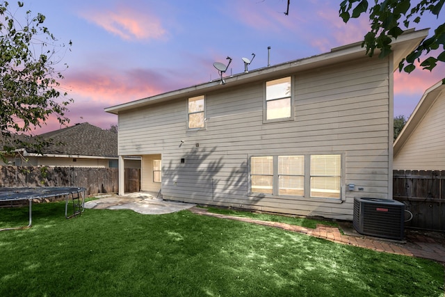 back house at dusk with cooling unit, a patio area, a trampoline, and a lawn