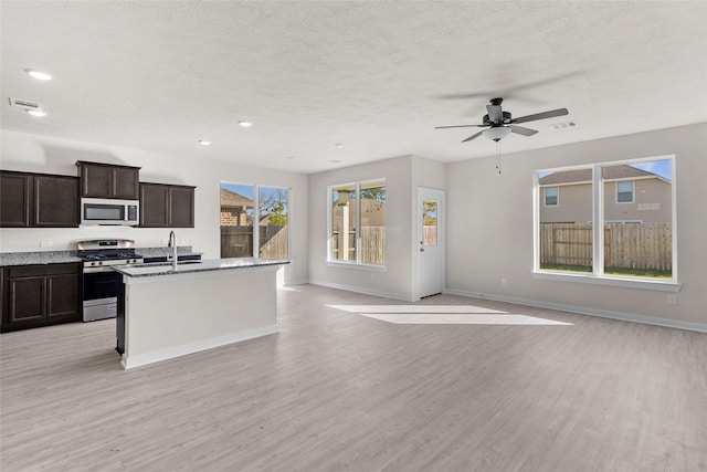 kitchen featuring ceiling fan, dark stone countertops, an island with sink, light hardwood / wood-style floors, and stainless steel appliances