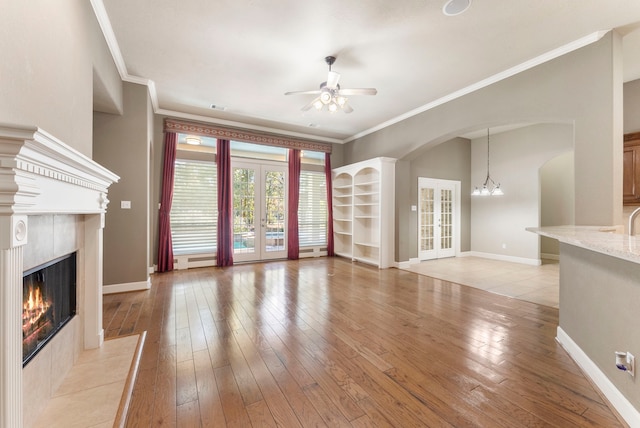 unfurnished living room featuring a tiled fireplace, ceiling fan with notable chandelier, french doors, and light wood-type flooring