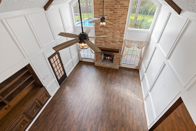 unfurnished living room with a brick fireplace, ceiling fan, dark wood-type flooring, beamed ceiling, and a high ceiling