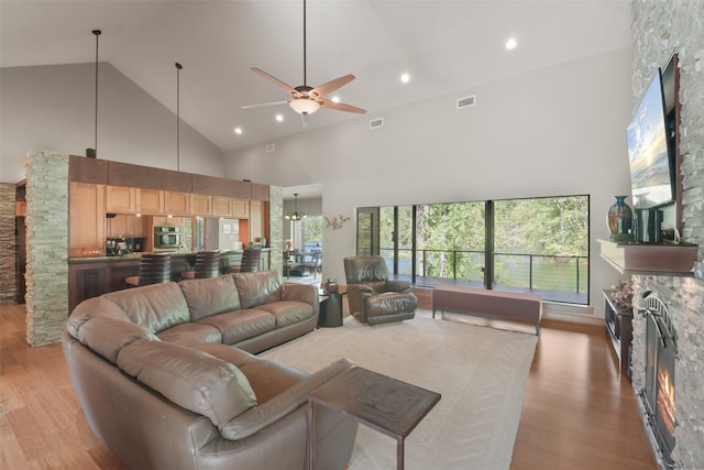 living room with a stone fireplace, high vaulted ceiling, light hardwood / wood-style floors, and ceiling fan with notable chandelier