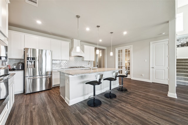 kitchen with hanging light fixtures, dark hardwood / wood-style flooring, premium range hood, a kitchen island with sink, and stainless steel appliances