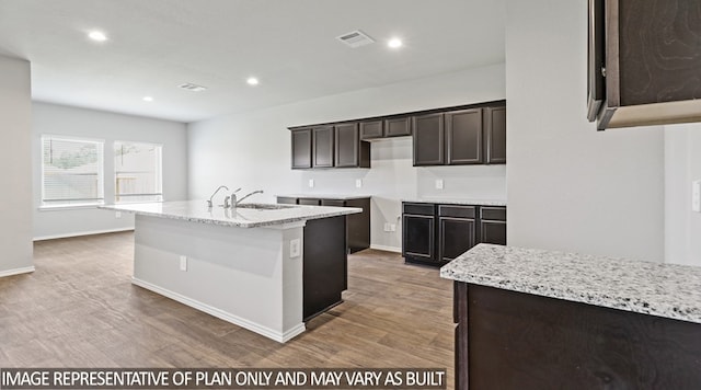 kitchen featuring light stone countertops, sink, dark brown cabinets, dark wood-type flooring, and a center island with sink