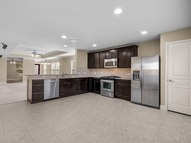 kitchen featuring kitchen peninsula, appliances with stainless steel finishes, dark brown cabinets, ceiling fan with notable chandelier, and a tray ceiling