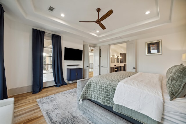 bedroom featuring ceiling fan, connected bathroom, light hardwood / wood-style flooring, and a tray ceiling