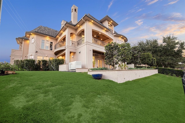 back house at dusk featuring a lawn and a balcony