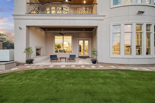 back house at dusk with exterior fireplace, a patio area, a balcony, and a lawn