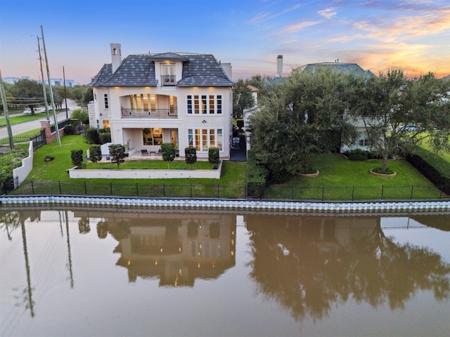 back house at dusk featuring a lawn, a balcony, and a water view