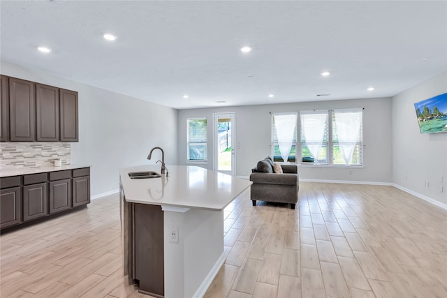 kitchen with sink, light wood-type flooring, dark brown cabinets, decorative backsplash, and a kitchen island with sink