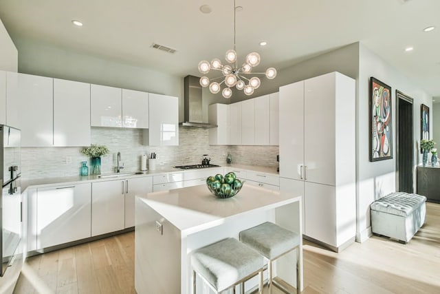 kitchen featuring wall chimney range hood, sink, white cabinetry, light hardwood / wood-style flooring, and a chandelier