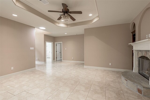 unfurnished living room featuring ornamental molding, ceiling fan, light tile patterned floors, a tray ceiling, and a stone fireplace