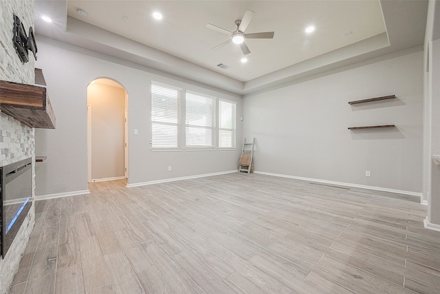 unfurnished living room featuring light hardwood / wood-style floors, a stone fireplace, a tray ceiling, and ceiling fan