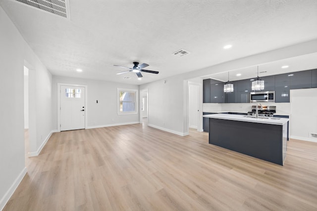 kitchen with appliances with stainless steel finishes, light wood-type flooring, a textured ceiling, decorative light fixtures, and a kitchen island with sink