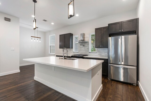 kitchen featuring wall chimney range hood, a center island with sink, sink, appliances with stainless steel finishes, and dark hardwood / wood-style flooring