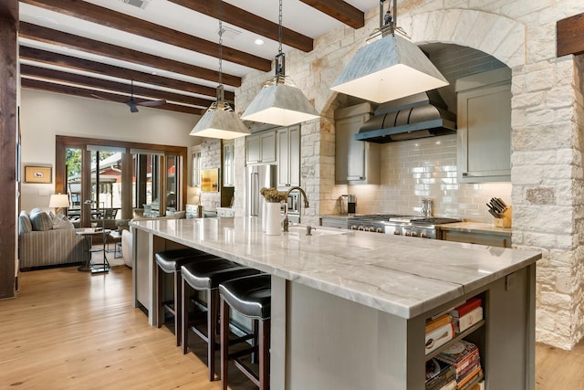 kitchen featuring beam ceiling, gray cabinets, an island with sink, and wall chimney exhaust hood