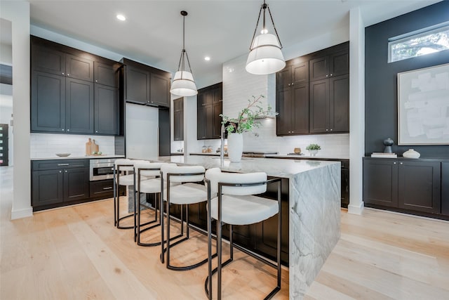 kitchen featuring light wood-type flooring, hanging light fixtures, a kitchen island with sink, and tasteful backsplash