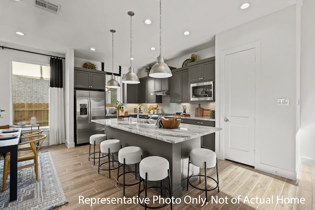 kitchen with a center island with sink, light stone counters, pendant lighting, light hardwood / wood-style floors, and stainless steel appliances