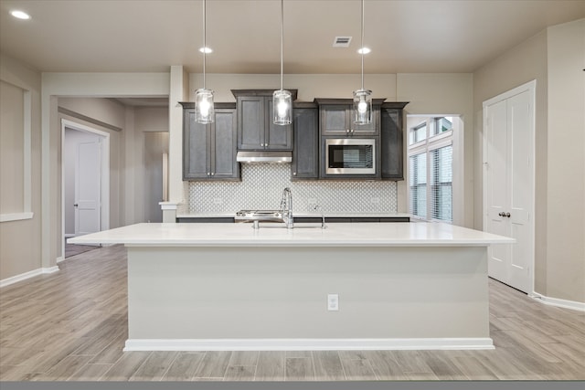 kitchen featuring stainless steel microwave, a kitchen island with sink, sink, light hardwood / wood-style floors, and decorative light fixtures