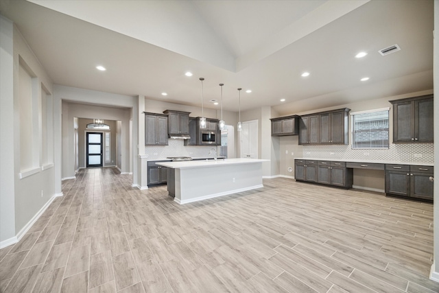 kitchen with hanging light fixtures, dark brown cabinets, a center island with sink, light hardwood / wood-style floors, and tasteful backsplash