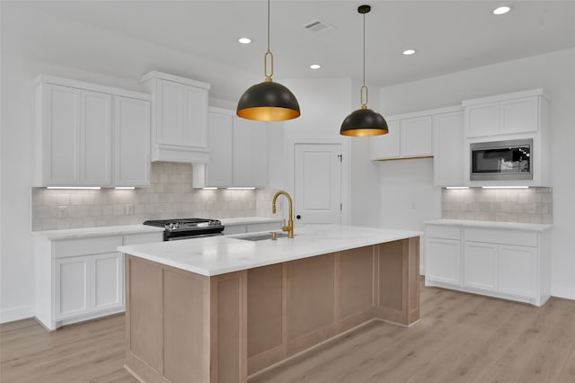 kitchen with white cabinets, hanging light fixtures, a center island with sink, built in microwave, and stainless steel gas range