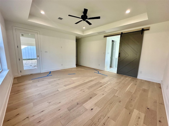 empty room with a raised ceiling, a barn door, ceiling fan, and light hardwood / wood-style flooring