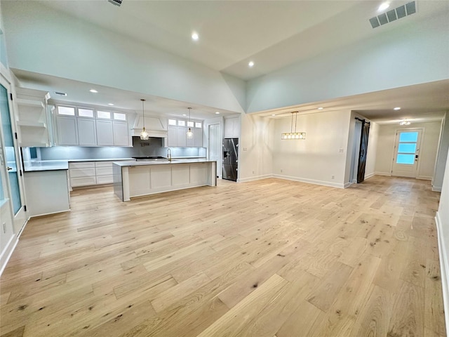 kitchen featuring stainless steel fridge with ice dispenser, light wood-type flooring, an island with sink, decorative light fixtures, and white cabinetry