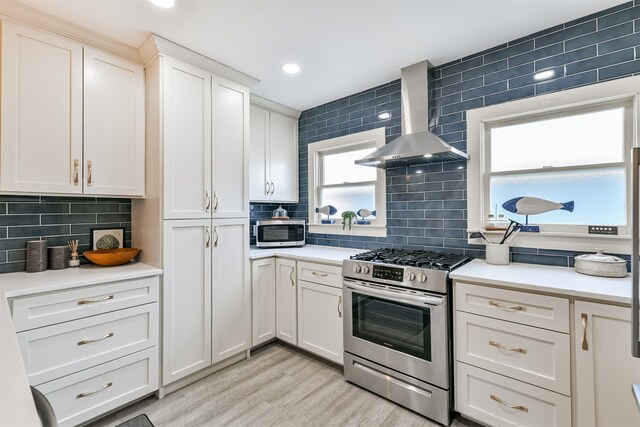 kitchen with decorative backsplash, wall chimney range hood, stainless steel appliances, white cabinets, and light hardwood / wood-style floors